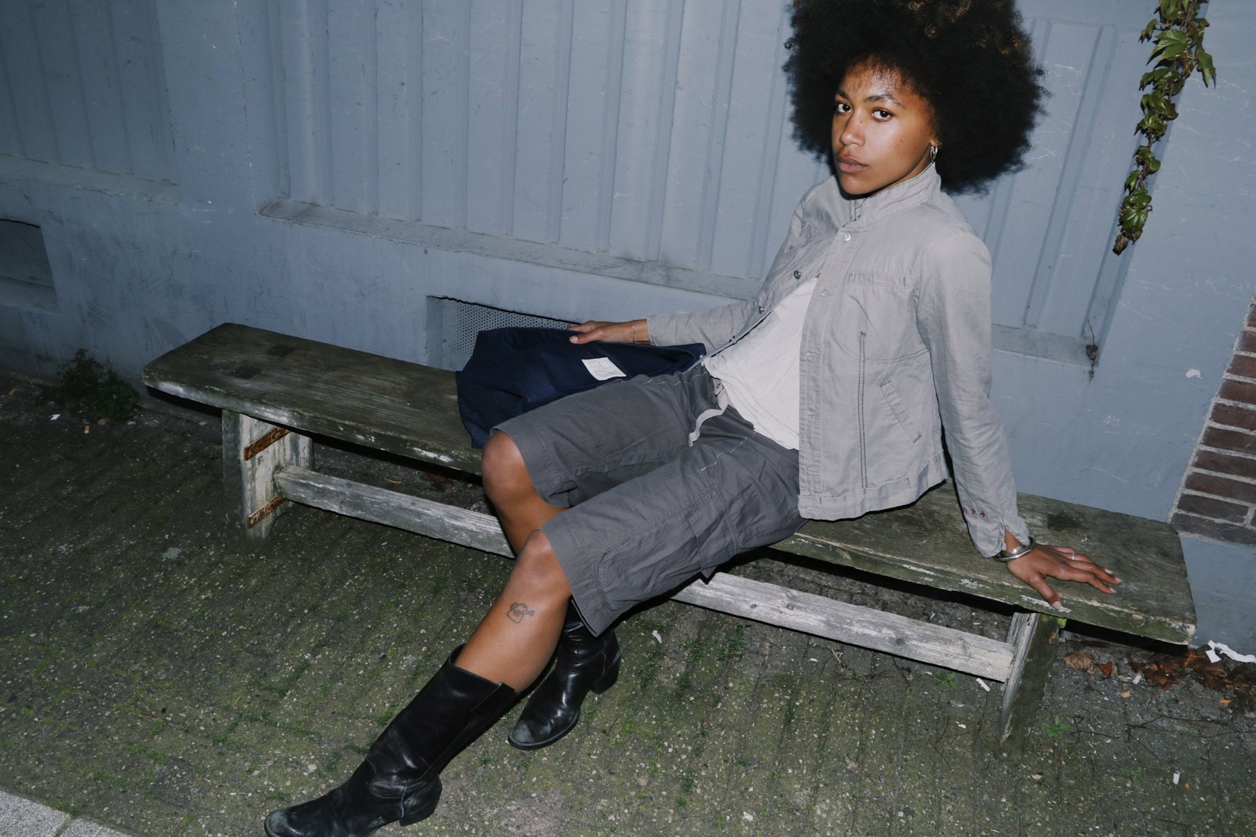 A friend in a grey jacket and grey cargo shorts leans back on a weathered bench at night, a navy canvas bag beside her.